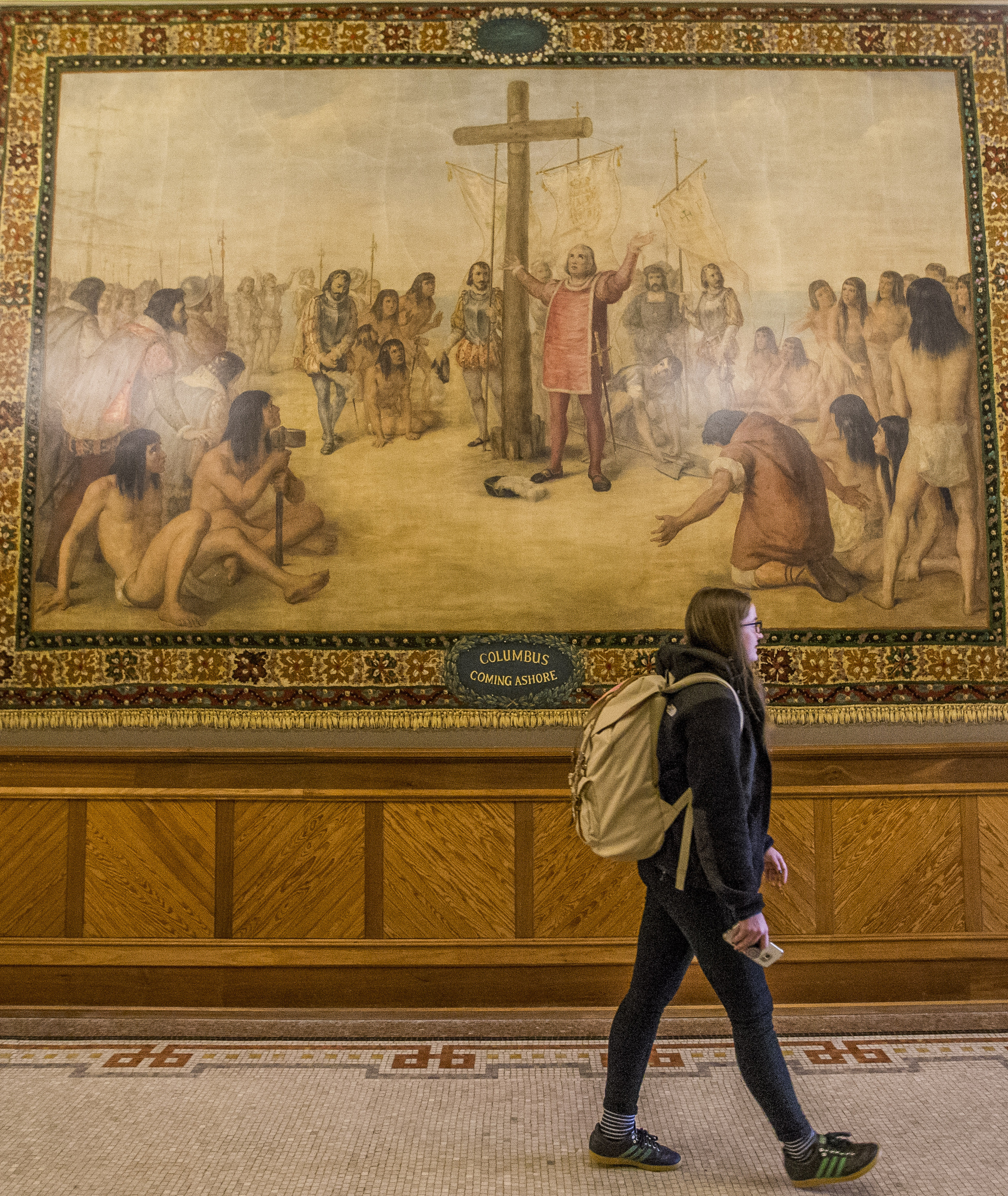 (Robert Franklin | South Bend Tribune via AP) In this Nov. 29, 2017, photo, Kristin Fabian walks by a mural of Christopher Columbus at Notre Dame in South Bend, Ind. The University of Notre Dame will cover murals in a campus building that depict Christopher Columbus in America, the school’s president said, following criticism that the images depict Native Americans in stereotypical submissive poses before white European explorers.